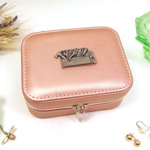Könnte beinhalten: Eine quadratische, rosafarbene Schmuckschatulle mit silbernem Reißverschluss und einem dekorativen Metall-Emblem mit Werkzeugen. Die Box ist geschlossen und steht auf einer weißen Oberfläche, mit Schmuck und dekorativen Gegenständen in der Nähe.