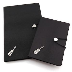 May include: Two black notebooks with silver violin charms. Each notebook has a button closure with an elastic band. The notebooks are different sizes, with a larger one on the left and a smaller one on the right.