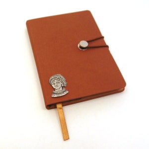 May include: A brown leather-bound journal with a silver bookmark featuring a portrait of a woman. The journal has a silver button closure and a brown ribbon.