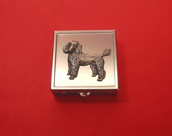 Puede incluir: Una caja de pastillas de metal plateado con un diseño de caniche negro en la parte delantera.