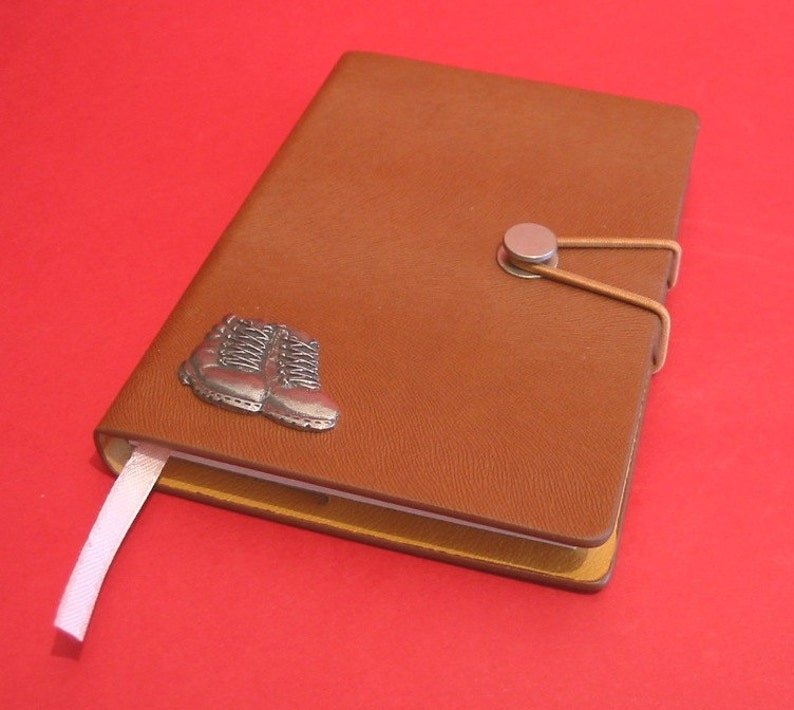 May include: A brown leather journal with a silver boot charm and a silver metal closure. The journal has a white ribbon bookmark.