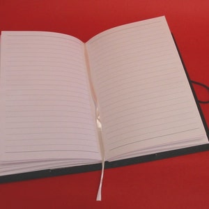 K&ouml;nnte beinhalten: Ein offenes Notizbuch mit linierten Seiten, einem schwarzen Einband und einem cremefarbenen Band als Lesezeichen. Die Seiten sind leer. Das Notizbuch liegt auf rotem Hintergrund.