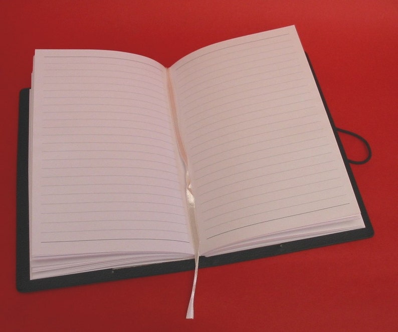 K&ouml;nnte beinhalten: Ein offenes Notizbuch mit linierten Seiten, einem schwarzen Einband und einem cremefarbenen Band als Lesezeichen. Die Seiten sind leer. Das Notizbuch liegt auf rotem Hintergrund.