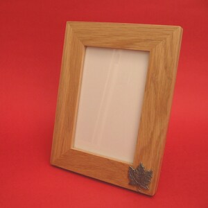 May include: A light oak picture frame with a silver maple leaf emblem in the corner. The frame is rectangular and angled, displaying a natural wood grain. The background is a solid red, highlighting the frame's design.