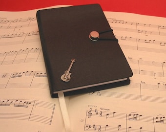 May include: A black leather bound journal with a silver guitar charm and a silver button closure. The journal is lying open on a sheet of music.
