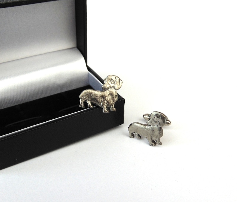 Puede incluir: Un par de gemelos de dachshund plateados en una caja de regalo negra. Los gemelos son detallados y realistas.