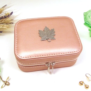 May include: A pink faux leather jewellery box with a silver maple leaf design on the lid. The box has a zipper closure and a silver pull tab.