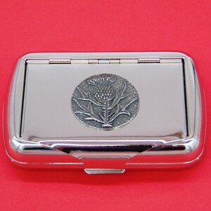 May include: A silver metal cigarette case with a hinged lid. The case features a raised, silver-toned emblem of a thistle flower.
