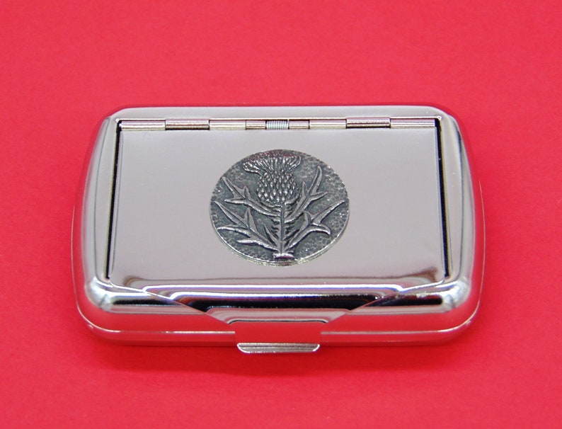 May include: A silver metal cigarette case with a hinged lid. The case features a raised, silver-toned emblem of a thistle flower.