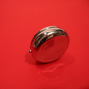 K&ouml;nnte beinhalten: Ein runder Beh&auml;lter aus silbernem Metall mit einem konzentrischen Kreisdesign auf dem Deckel. Der Beh&auml;lter befindet sich auf einem roten Hintergrund.