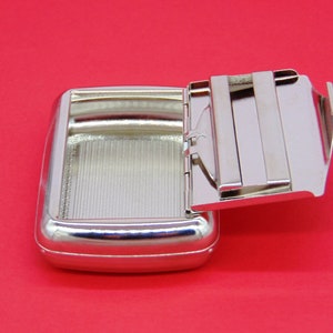 May include: A silver metal cigarette case with a hinged lid. The lid is open and the case is empty.
