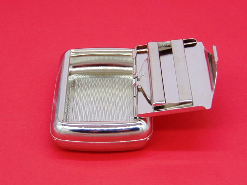 May include: A silver metal cigarette case with a hinged lid. The lid is open and the case is empty.