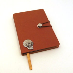May include: A brown leather journal with a silver skull charm and a silver button closure. The journal has a brown ribbon bookmark.