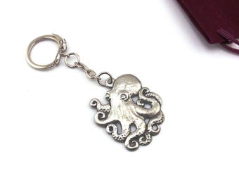 May include: A silver-colored octopus keychain with a detailed design. The octopus charm is attached to a chain and keyring. A maroon drawstring bag is in the background.