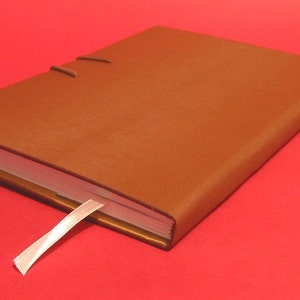 May include: A brown leather-bound journal with a white ribbon bookmark. The journal is closed and lying on a red surface.