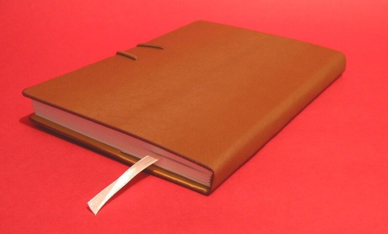 May include: A brown leather-bound journal with a white ribbon bookmark. The journal is closed and lying on a red surface.