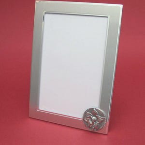 May include: A rectangular silver-colored picture frame with a decorative floral emblem on the lower right corner. The frame has a clean, modern design and a white interior. The background is a solid red.