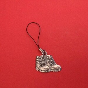 May include: A silver-toned metal charm shaped like a pair of hiking boots, featuring detailed laces and tread patterns. The charm is attached to a thin, black cord loop, designed for attachment to small items like mobile phones or keychains.