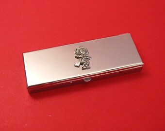 May include: A rectangular, silver-colored business card holder with a small, decorative figure on the top. The figure appears to be a stylized person. The background is a solid red color.