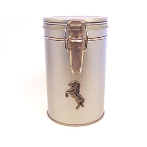 May include: A silver metal canister with a gold latch and a black horse silhouette on the front.
