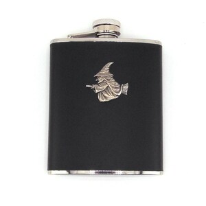 May include: A black stainless steel hip flask with a silver witch emblem. The flask has a silver cap and trim. The witch is depicted riding a broom. The flask is rectangular and has a smooth finish.
