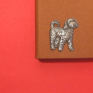 K&ouml;nnte beinhalten: Eine silberne Hundefigur auf braunem Lederhintergrund. Der Hund steht auf allen vieren und hat seinen Schwanz eingerollt.