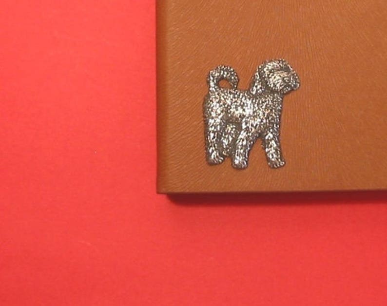 K&ouml;nnte beinhalten: Eine silberne Hundefigur auf braunem Lederhintergrund. Der Hund steht auf allen vieren und hat seinen Schwanz eingerollt.
