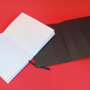 Puede incluir: Un cuaderno blanco con l&iacute;neas y una cubierta negra con un marcador de cinta negro. El cuaderno est&aacute; abierto y la cubierta est&aacute; cerrada.