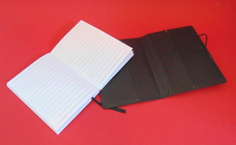 Puede incluir: Un cuaderno blanco con l&iacute;neas y una cubierta negra con un marcador de cinta negro. El cuaderno est&aacute; abierto y la cubierta est&aacute; cerrada.