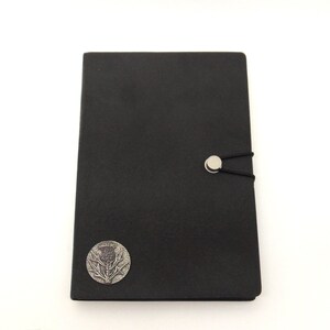 May include: Black leather journal with a silver thistle emblem and a silver button closure. The journal has an elastic band closure.