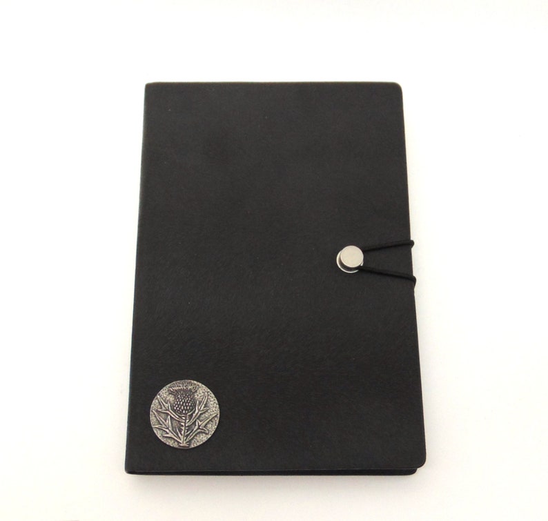 May include: Black leather journal with a silver thistle emblem and a silver button closure. The journal has an elastic band closure.