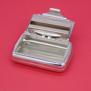 May include: A silver metal cigarette rolling tray with a hinged lid. The tray has a grid pattern on the bottom and a slot for holding rolling papers.