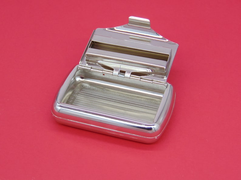 May include: A silver metal cigarette rolling tray with a hinged lid. The tray has a grid pattern on the bottom and a slot for holding rolling papers.