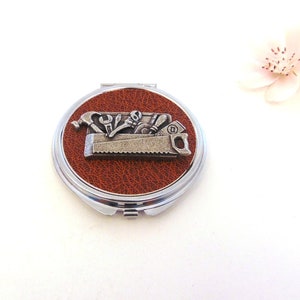 May include: A compact mirror with a brown leatherette cover and a silver metal trim. The mirror features a pewter design of a toolbox with tools, including a hammer, saw, wrench, and screwdriver.