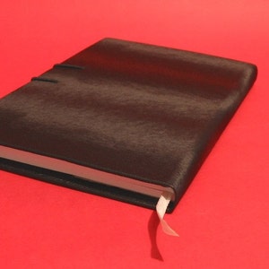 K&ouml;nnte beinhalten: Ein geschlossenes, schwarz ledergebundenes Notizbuch mit strukturierter Oberfl&auml;che. Das Notizbuch hat ein wei&szlig;es Band-Lesezeichen und ist vor einem roten Hintergrund platziert. Der Einband wird mit einem schwarzen Gummiband gesichert.
