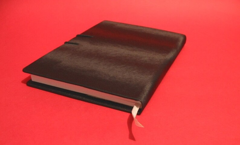 K&ouml;nnte beinhalten: Ein geschlossenes, schwarz ledergebundenes Notizbuch mit strukturierter Oberfl&auml;che. Das Notizbuch hat ein wei&szlig;es Band-Lesezeichen und ist vor einem roten Hintergrund platziert. Der Einband wird mit einem schwarzen Gummiband gesichert.