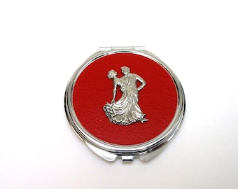 Puede incluir: Un espejo compacto redondo plateado con un centro rojo con aspecto de cuero. Un relieve plateado de una pareja bailando está centrado en el fondo rojo. El compacto tiene una apertura con bisagras y un borde metálico brillante.