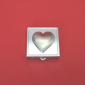 May include: A silver heart-shaped object sits within a square, silver-toned box with a hinged clasp. The heart has a gradient effect, lighter at the base and darker at the top. The background is a solid red colour.