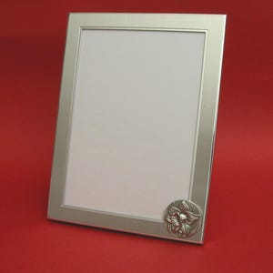 May include: Silver rectangular picture frame with a floral emblem in the lower right corner. The frame has a clean, modern design and a white interior, set against a red background.