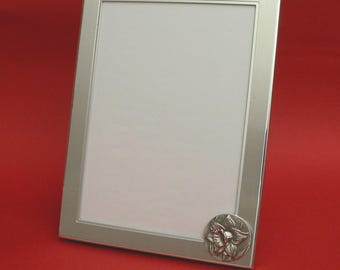 Puede incluir: Marco de fotos rectangular plateado con un emblema floral en la esquina inferior derecha. El marco tiene un diseño limpio y un interior blanco, sobre un fondo rojo.