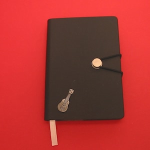 May include: A black leather journal with a silver ukulele charm and a silver button closure. The journal has a white ribbon bookmark.