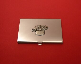 Puede incluir: Un porta tarjetas de visita de metal plateado con un diseño de gorro de chef en relieve.