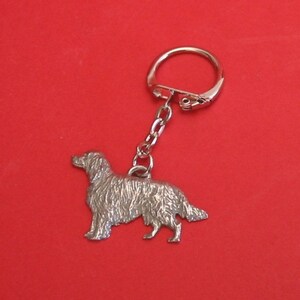 Pode incluir: Um chaveiro de prata com um amuleto de cão golden retriever detalhado. O amuleto é preso a um anel partido de prata e uma corrente de prata.