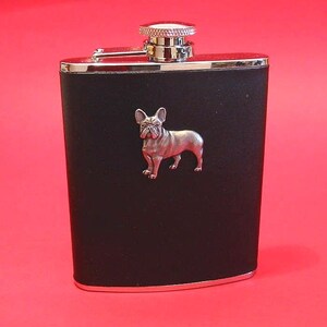 May include: A black rectangular hip flask featuring a silver-toned metal French Bulldog emblem centred on the front. The flask has a polished silver-toned metal top with a screw-on cap and a hinged arm, providing a secure closure for the container.