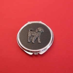 May include: A silver compact mirror with a black leatherette cover featuring a silver-toned silhouette of a dog.
