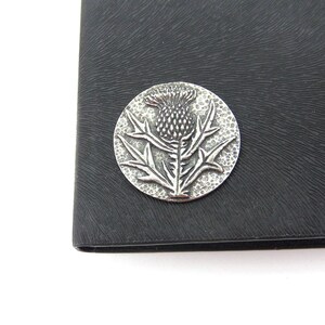 May include: A silver-toned metal pin featuring a raised design of a thistle flower. The pin is circular and has a textured surface.