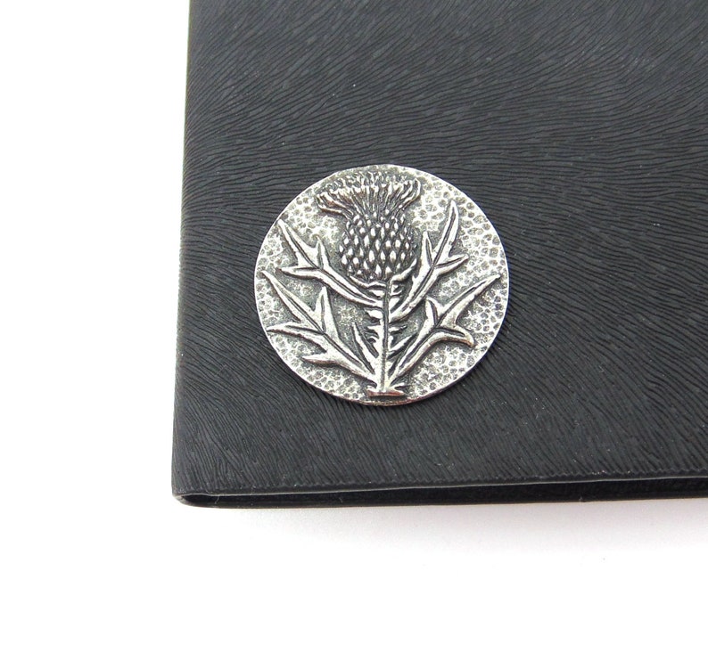 May include: A silver-toned metal pin featuring a raised design of a thistle flower. The pin is circular and has a textured surface.