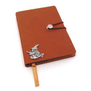 Könnte beinhalten: Ein braunes, ledergebundenes Tagebuch mit einer silbernen Hexe auf dem vorderen Umschlag. Das Tagebuch hat ein Lesezeichen aus Band und einen silbernen Knopfverschluss.