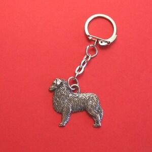 May include: A silver keychain featuring a detailed miniature of an Australian Shepherd dog. The dog is standing with its head turned to the side, and its tail is curled up. The keychain is attached to a silver ring and chain.