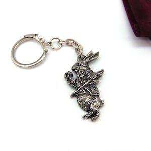 May include: A silver keychain featuring a detailed depiction of the White Rabbit from Alice in Wonderland, holding a pocket watch and running. The keychain has a silver ring and chain.